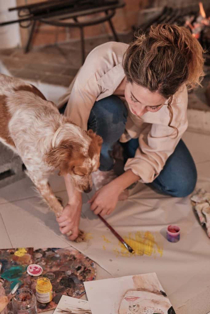 Creative female artist bonding with pet using paintbrush and paw print while drawing on fabric in modern studio