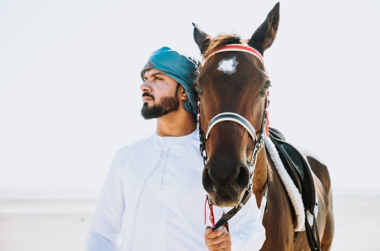 Young adult with Kandura, the emirates traditional clothes, riding his horse in the desert