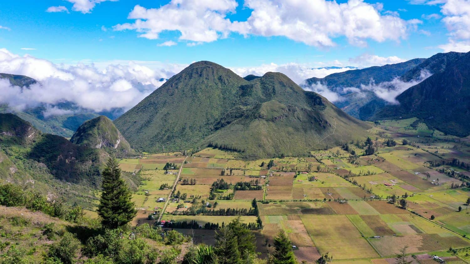 Aerial view of the Pululahua Crater in Ecuador showing the large volcanic dome above cultivated fields and pastures, with cloud‐topped hills and scattered houses. The mist ready to fills the caldera.