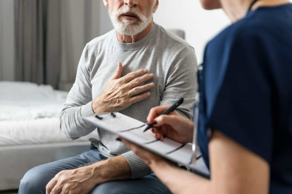 Patient complains of chest pain, heart problems to nurse. GP in blue uniform talk to elderly man, provide professional consultation, writing notes in medical records about disease and treatment plan