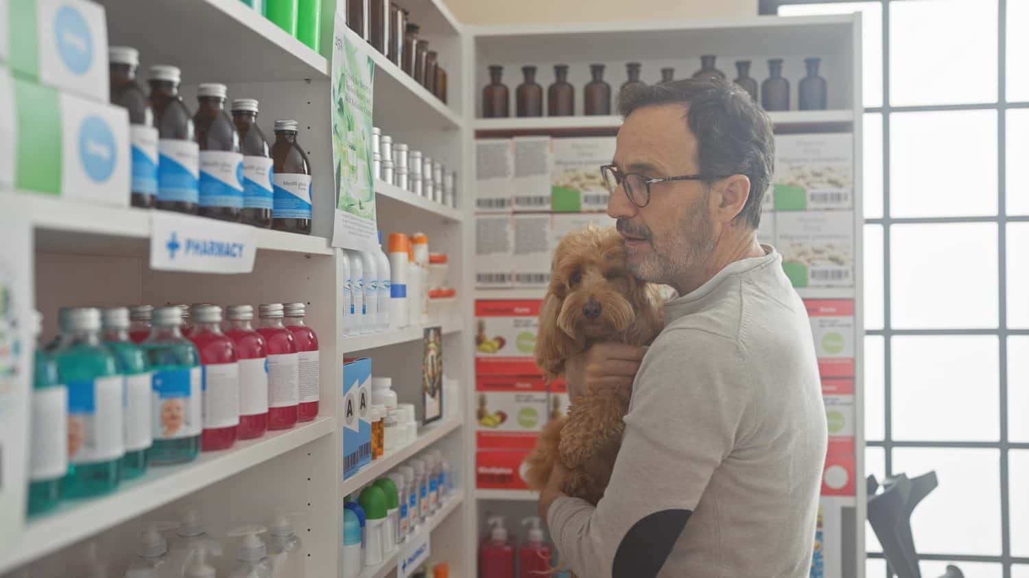 A middle-aged hispanic man affectionately holding his poodle inside a well-stocked pharmacy