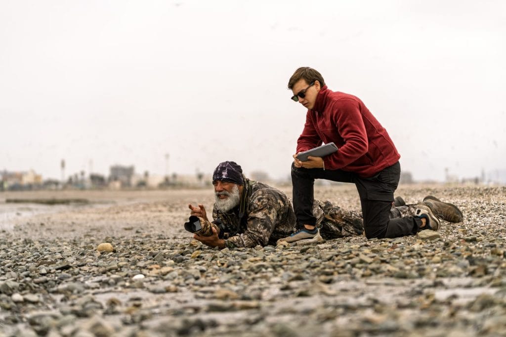 Two men observing birds and nature, one capturing wildlife with a camera, the other recording data on a tablet