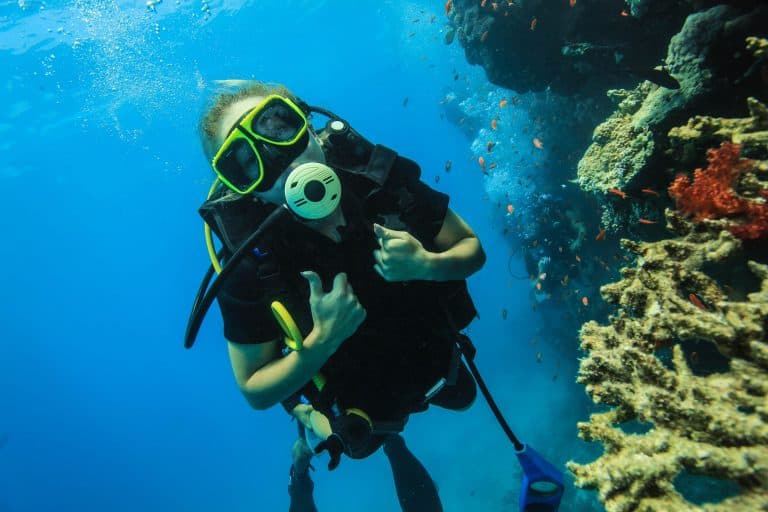 Scuba diver underwater making OK hand sign near vibrant coral reef with colorful tropical fish. Underwater adventure, marine life, and diving experience concept.