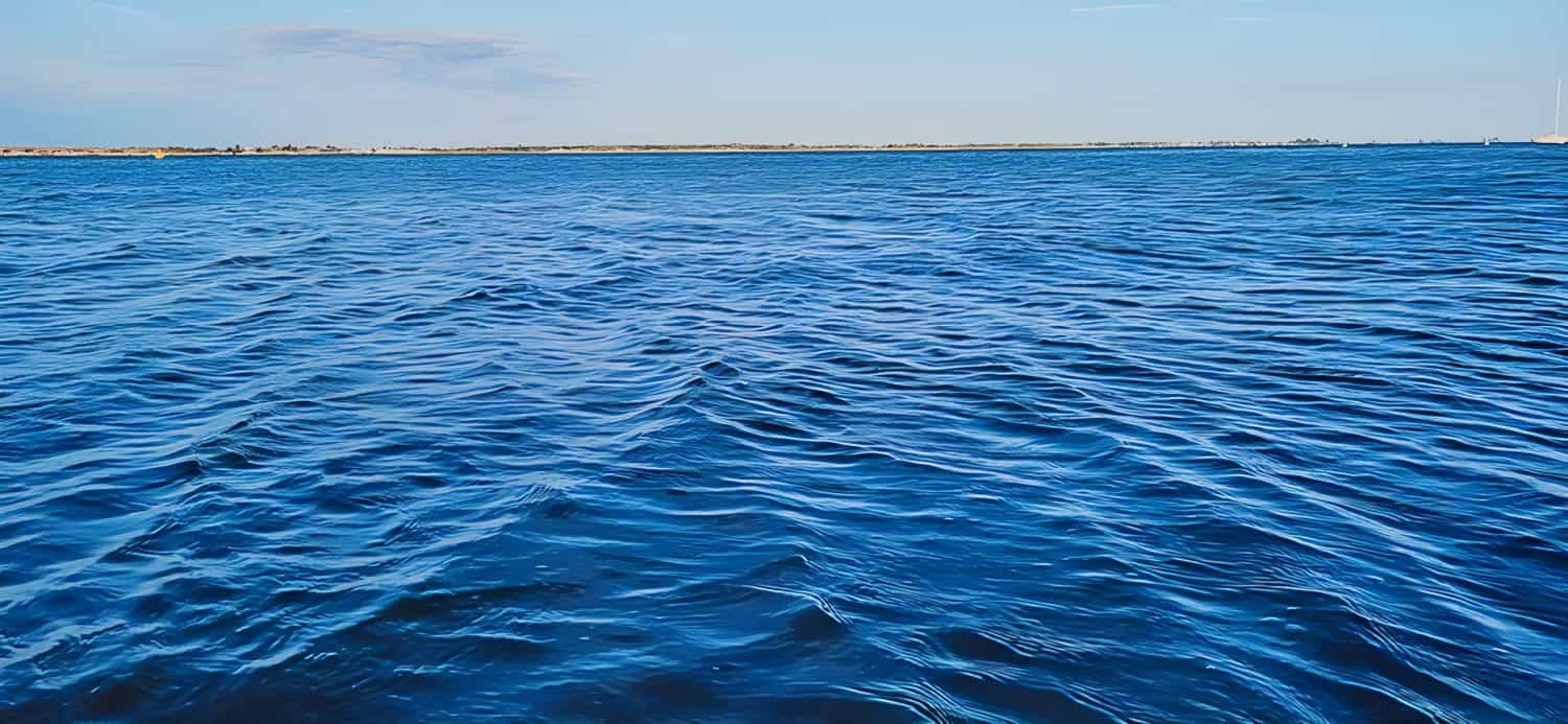 A peaceful expanse of deep blue water, rippled by a gentle breeze. On the horizon, a thin strip of land and a solitary sailboat evoke freedom and vastness. The clear sky enhances the serenity of this ocean scene.