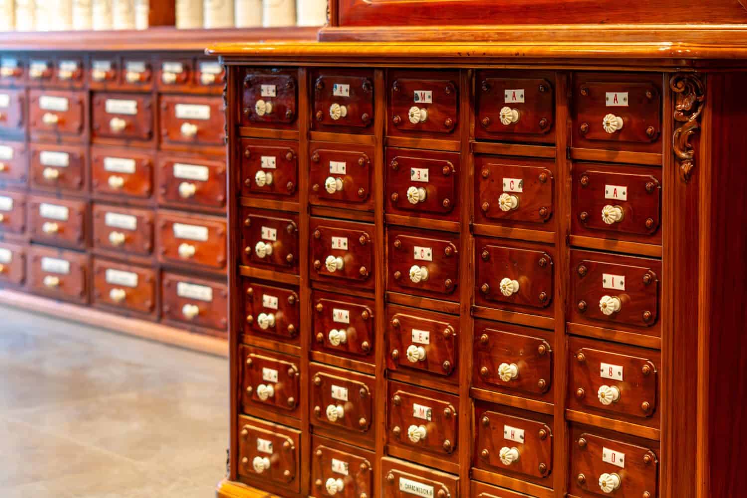 Vintage wooden apothecary cabinet featuring multiple small drawers with white ceramic knobs and letter labels, blurred second cabinet in the background, warm interior lighting, side perspective, selec