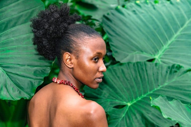 Young African woman in profile without make up with beans necklace and bare back in tropical jungle or forest on background of big green palm leaves at daytime. Cultures, traditions and travel concept