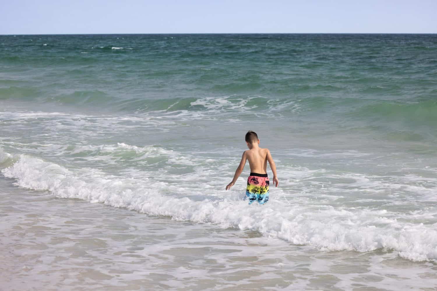 A child stands in the water at Gulf Shores Alabama beach. Ocean waves wash over small feet as sunlight brightens the scene. Families enjoy the beach day during summer.