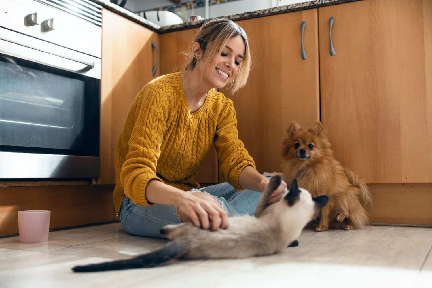 Shot of beautiful young woman playing with her cute lovely animals sitting on the floor in the kitchen at home.